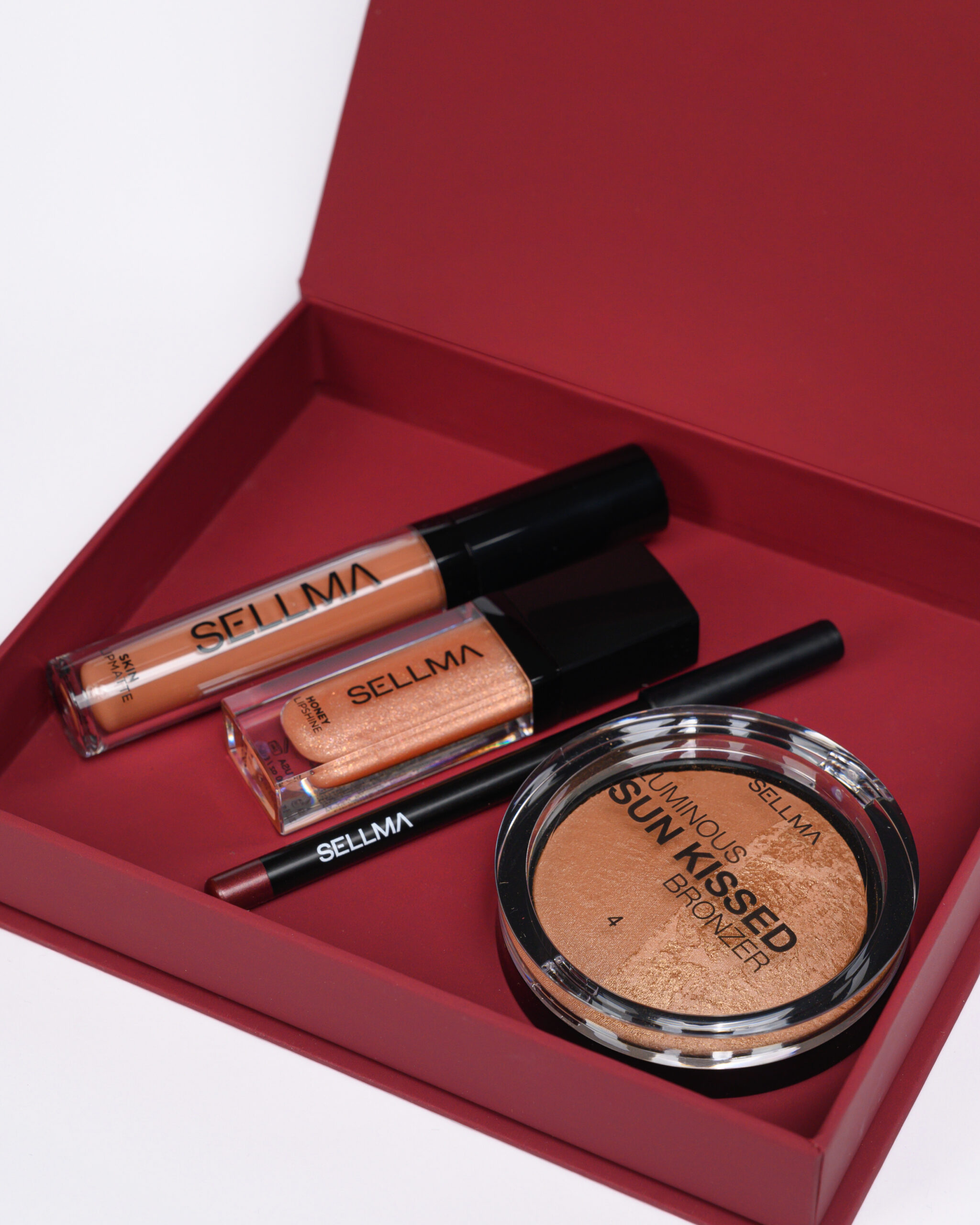 Open red makeup box with multiple nude lip gloss tubes labeled SELLMA and a round bronzer compact reading Sun-Kissed Bronzer in the foreground.